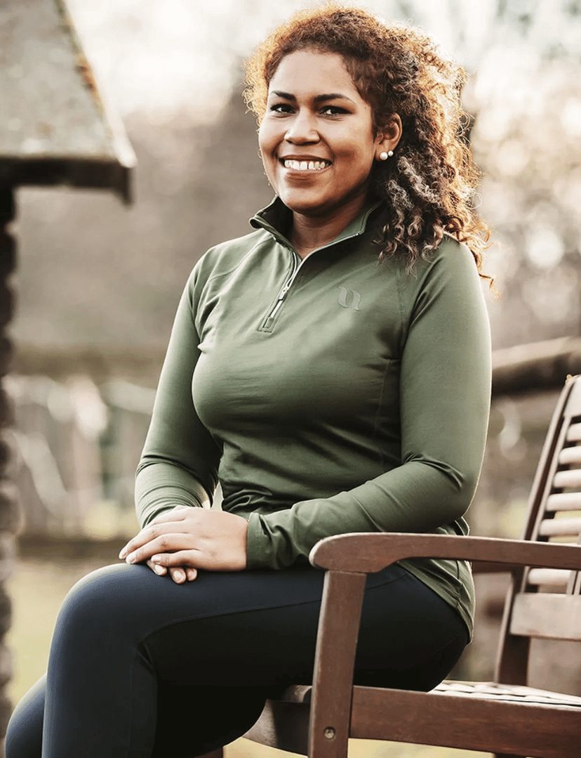 A smiling rider in a green Back on Track Leia P4G long sleeve top sitting on a bench by the barn