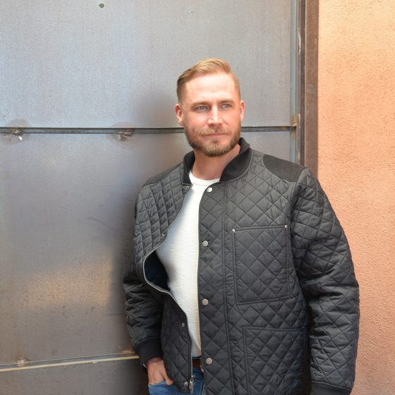 A man in an olive Chestnut Bay Heritage quilted barn jacket standing by a metal barn wall