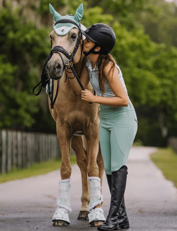 A rider in a Dapplebay Dragonfly Meadow top and green breeches kissing her palomino pony in a matching ear bonnet and boots