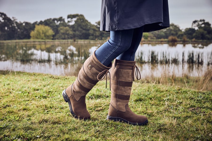 A woman in brown Dublin River Boots IV walking across a grassy field by a lake