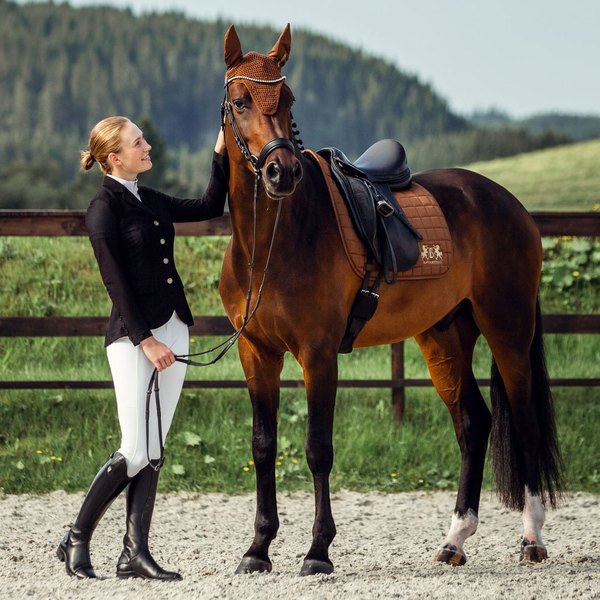 A rider in a black B Vertigo Gabrielle mesh show jacket and white breeches standing beside her bay horse in a green paddock