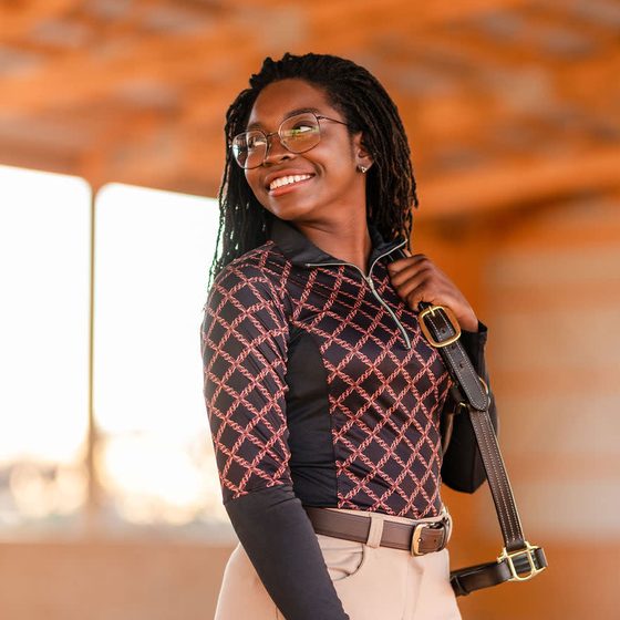 A smiling rider in an Equine Couture patterned riding top and Heather full seat breeches in an indoor arena