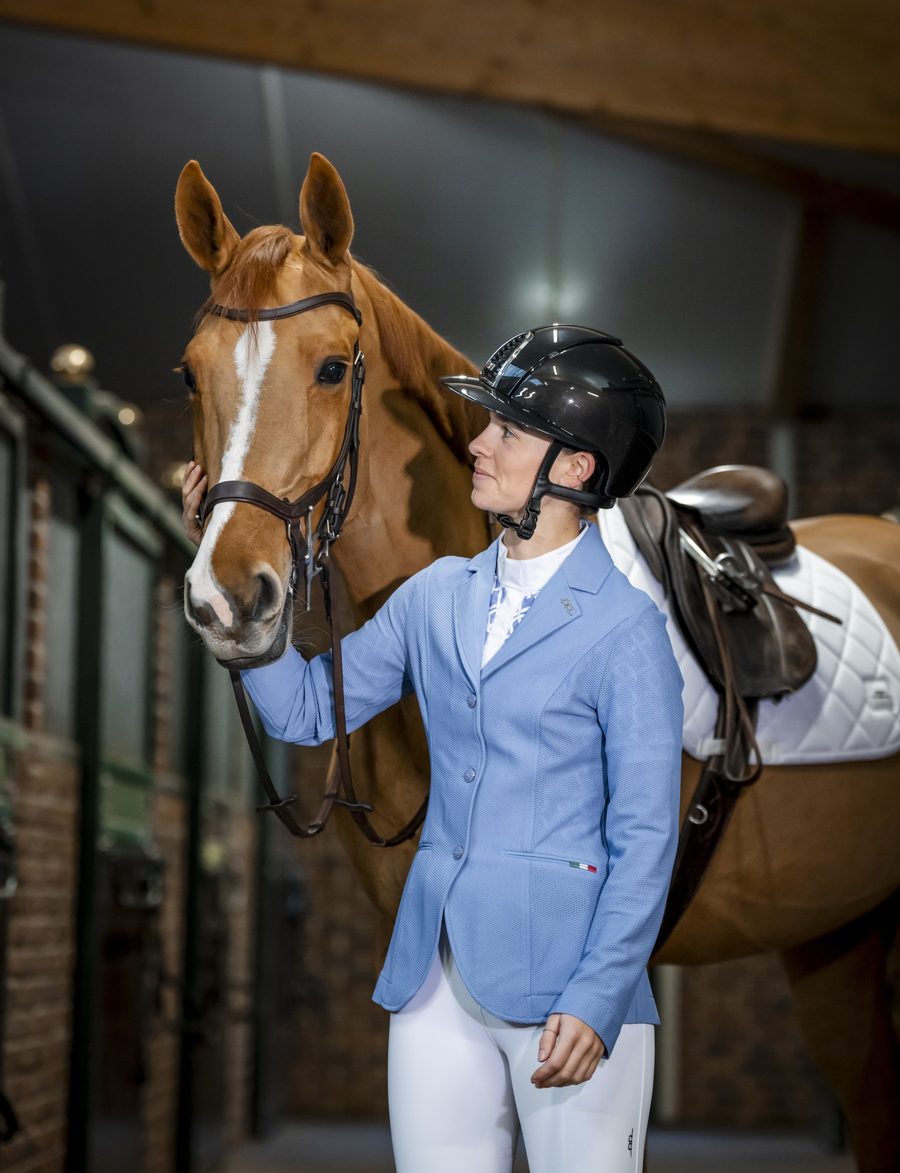 A rider in a Sicilian blue Alessandro Albanese Motion Lite show coat holding her chestnut horse in the barn aisle