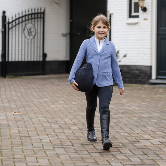 A young rider in a light blue Alessandro Albanese show coat walking across a cobbled stable yard with her helmet