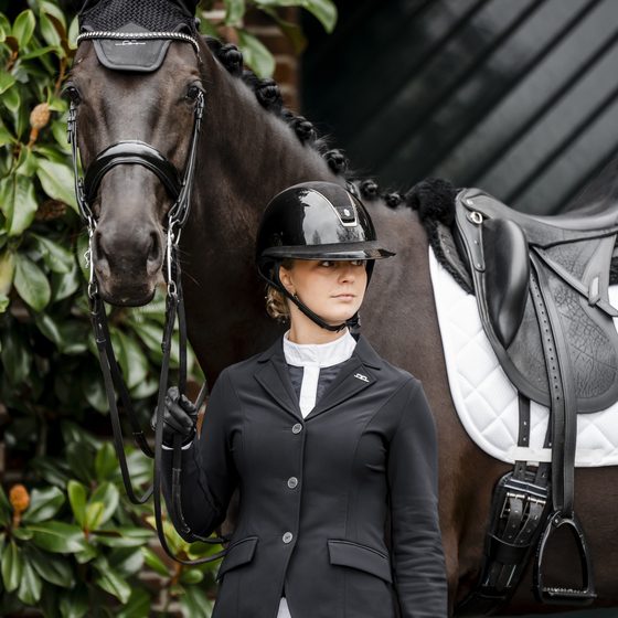 A rider in a black Alessandro Albanese show coat standing beside her bay horse