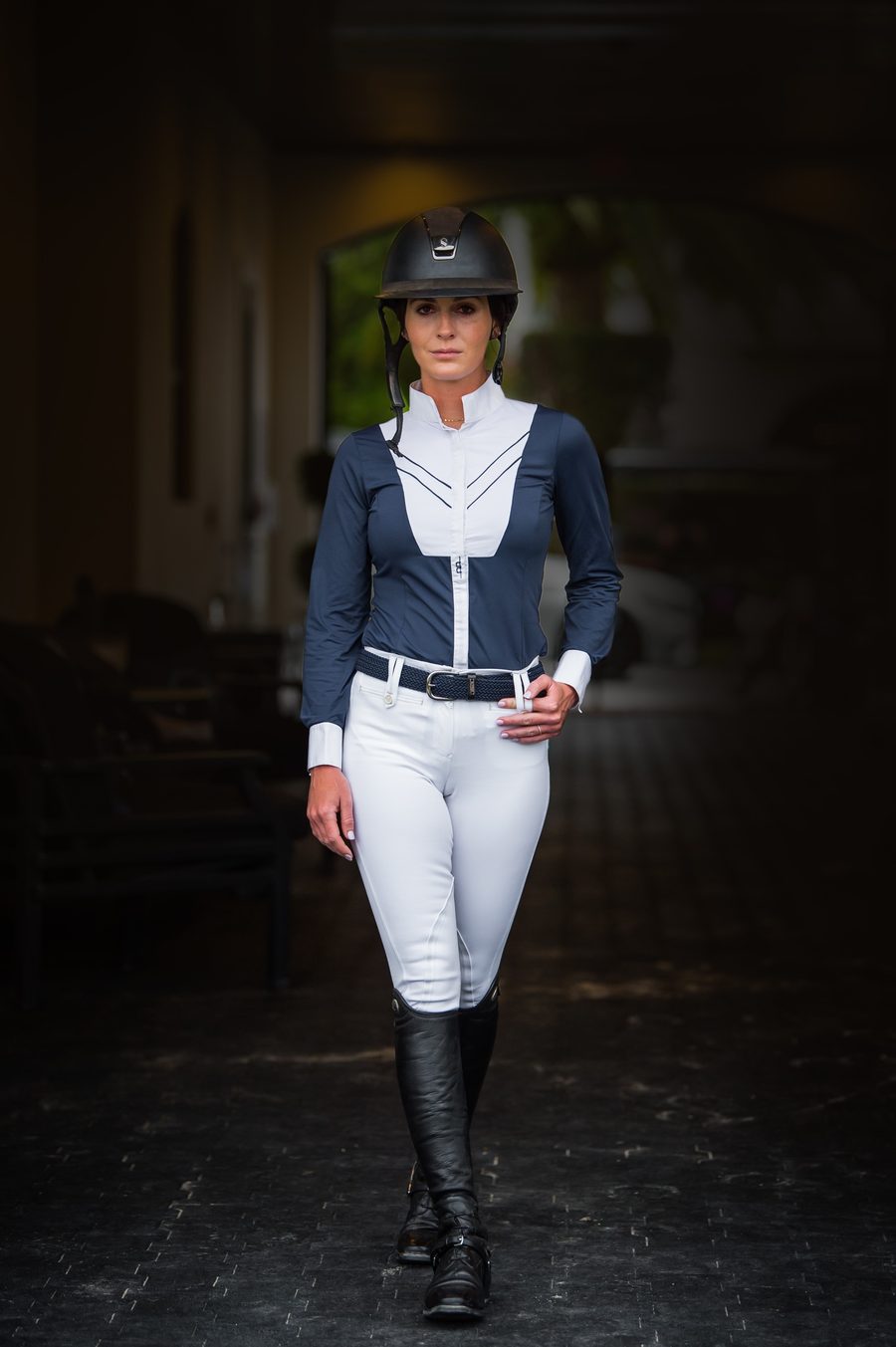 A rider in white Alessandro Albanese Athens breeches, a navy and white show shirt and tall boots walking down the barn aisle