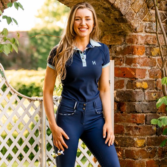 A smiling rider in a navy Horseware Newmarket polo and riding tights under a garden archway