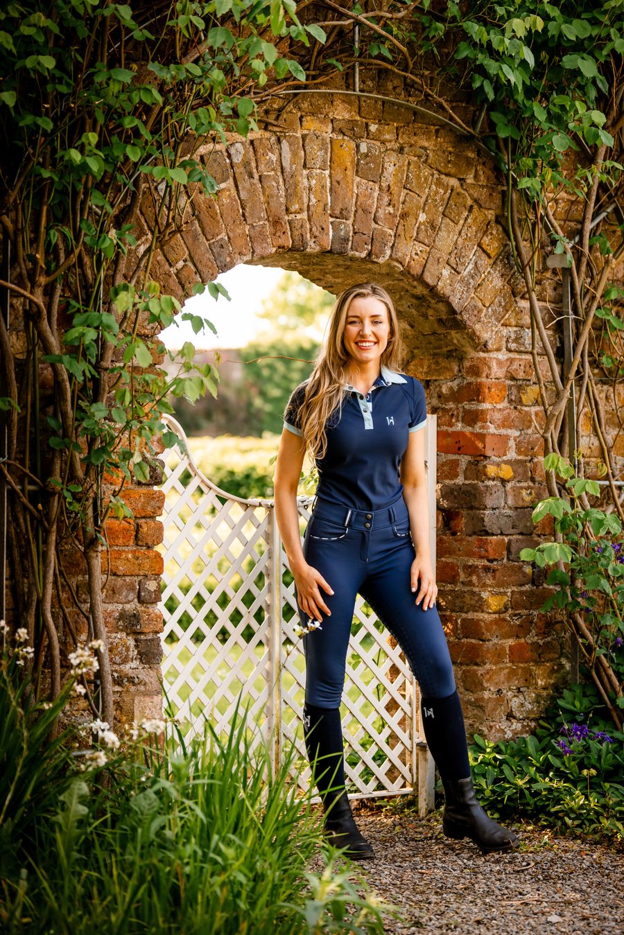 A smiling rider in a Horseware Newmarket polo and riding tights under a garden archway