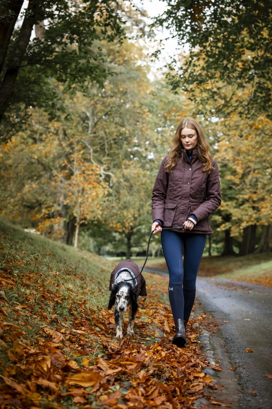 A rider in a Horseware Newmarket wax jacket walking her dog down an autumn lane