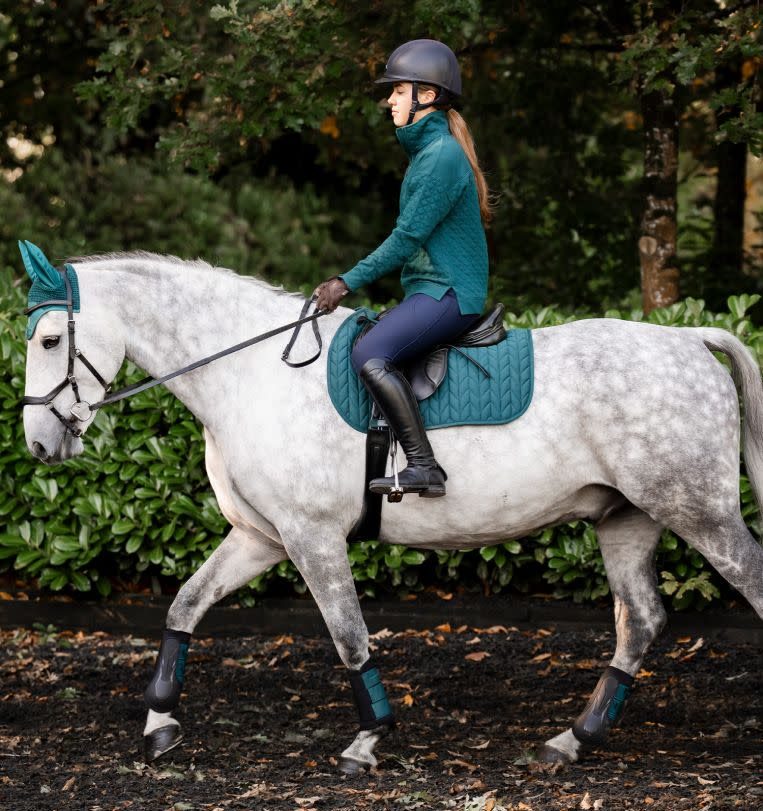 A rider in a teal jacket on a grey horse wearing a matching Horseware Close Contact saddle pad and boots