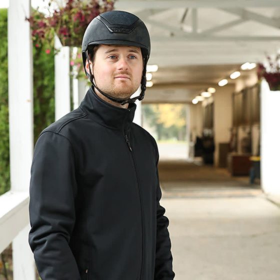 A rider in a Kerrits Men's Softshell Riding Jacket and helmet in a barn aisle