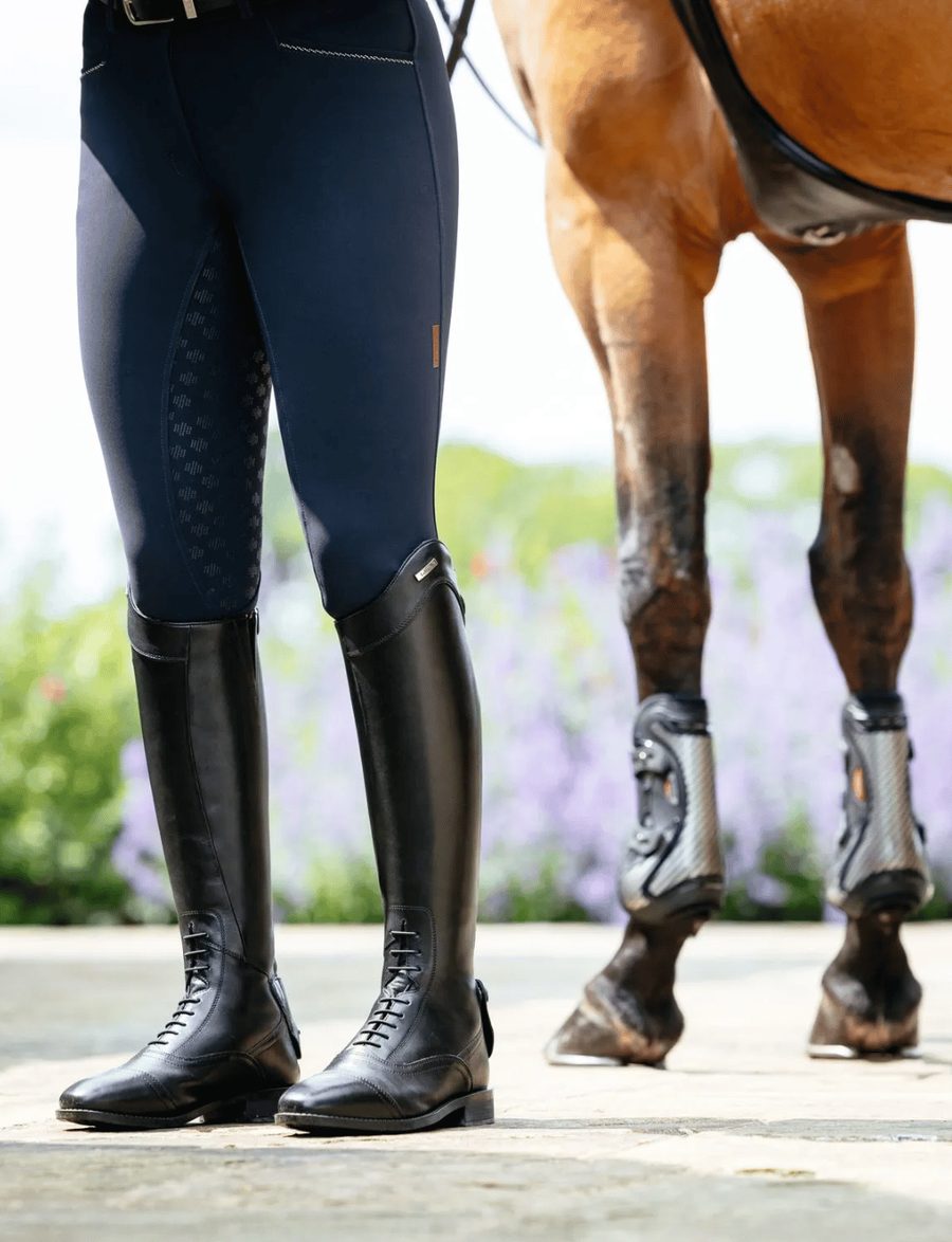 A rider in black LeMieux field riding boots and navy breeches standing beside her horse