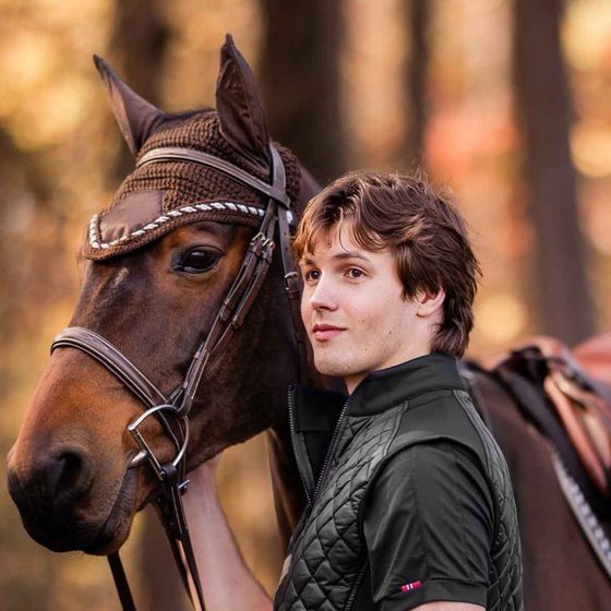 A young rider in a quilted vest beside his bay horse