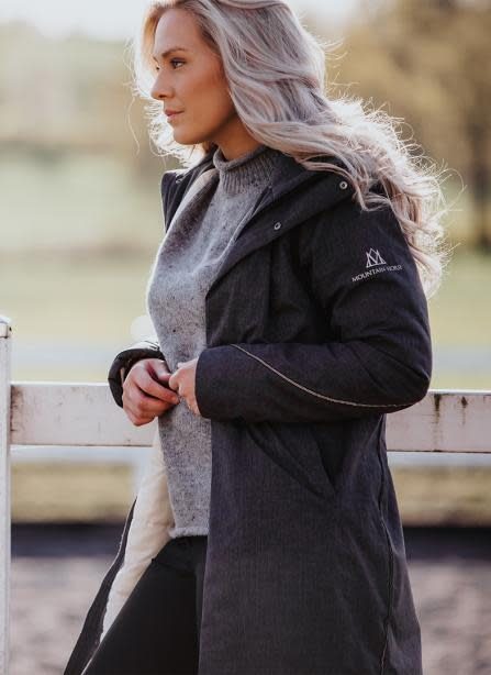 A rider in a long black Mountain Horse Alicia coat leaning on an arena fence