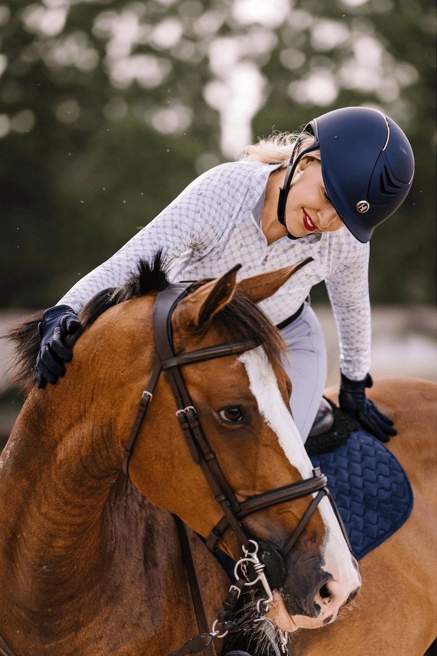 A smiling rider in a navy One K helmet leaning down to hug her chestnut horse's neck