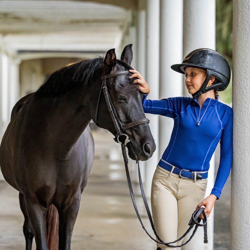 A young rider in a blue Ovation sun shirt and tan breeches with her dark bay horse under a colonnade