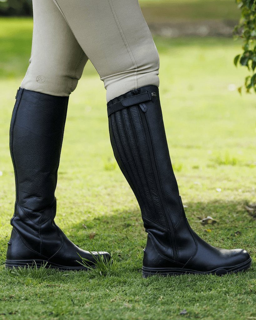 A rider walking across a lawn in black Ovation Moorland II waterproof tall boots and tan breeches