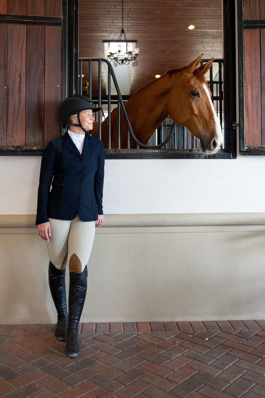 A rider in a navy RJ Classics show coat leaning on the barn wall below her chestnut horse looking out of his stall window