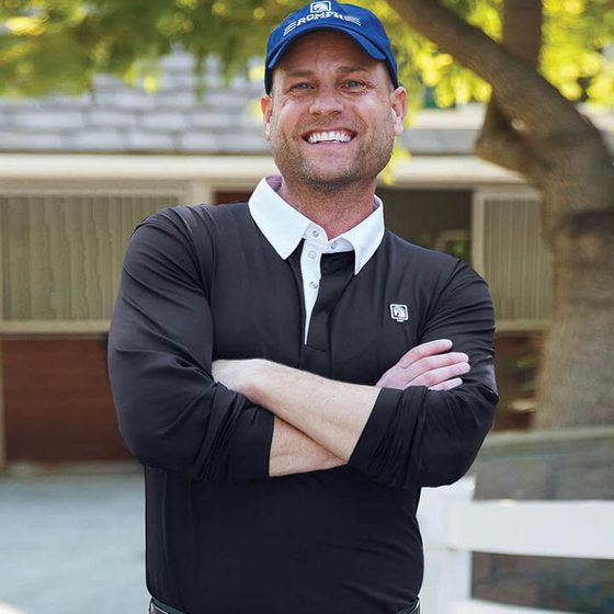 A smiling rider in a navy Romfh cap and black top outside a barn