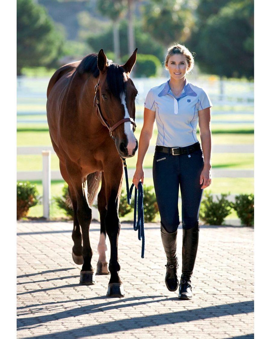 A rider in navy Romfh Sarafina knee patch breeches and a white sun shirt leading her bay horse
