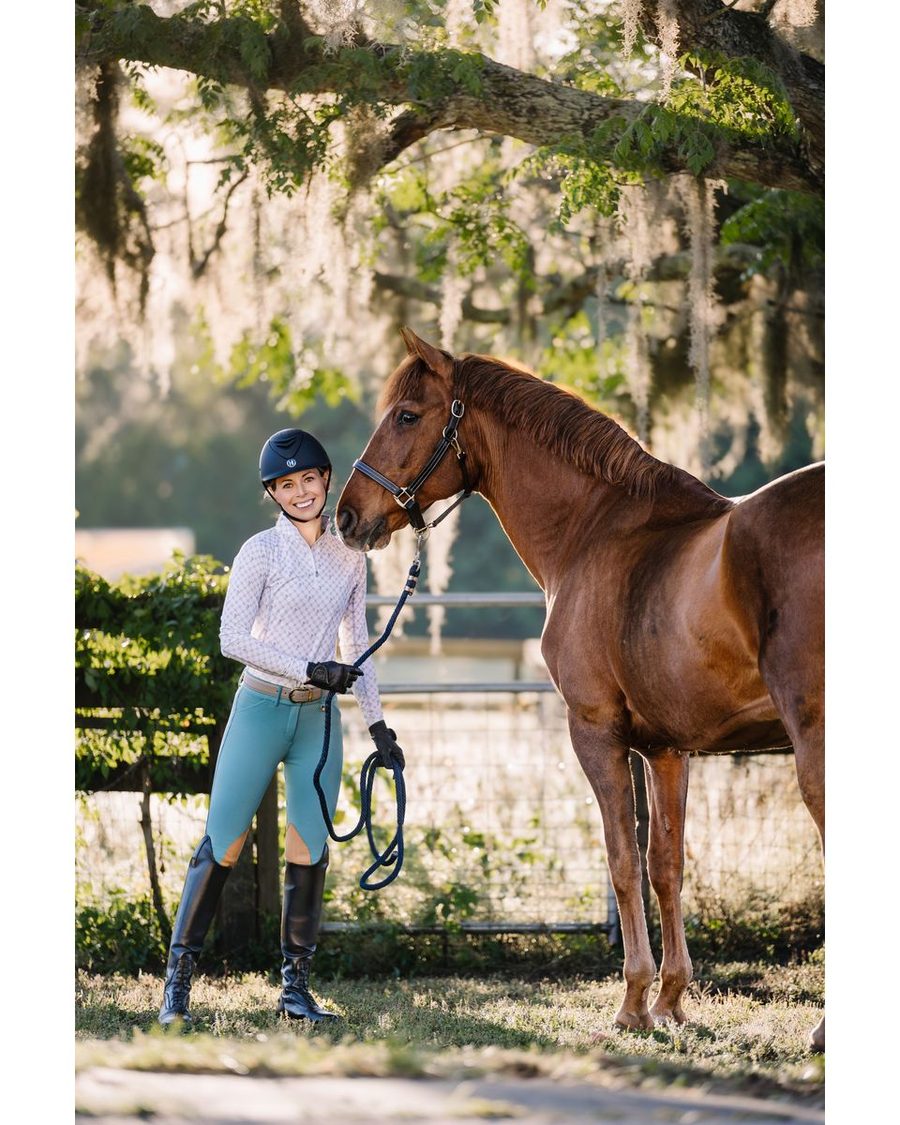 A rider in teal Romfh full seat breeches standing with her chestnut horse under the trees