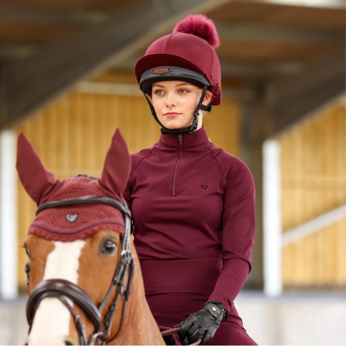A chestnut horse in a burgundy ear bonnet beside a rider in a burgundy hat and top