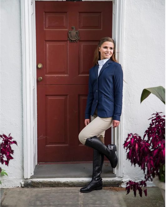 A rider in a navy show coat, tan breeches and black Shires Moretta tall boots outside a red door