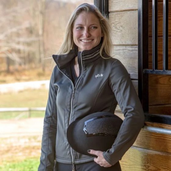 A smiling rider in a TuffRider jacket holding her helmet by the barn
