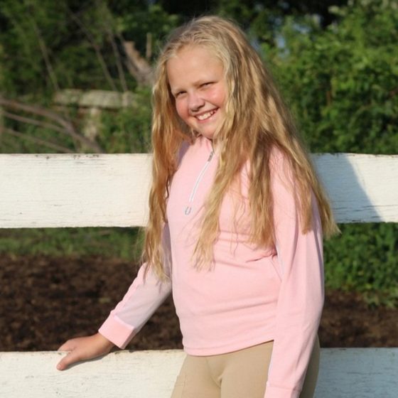A smiling girl in a pink TuffRider shirt and tan breeches by a white fence