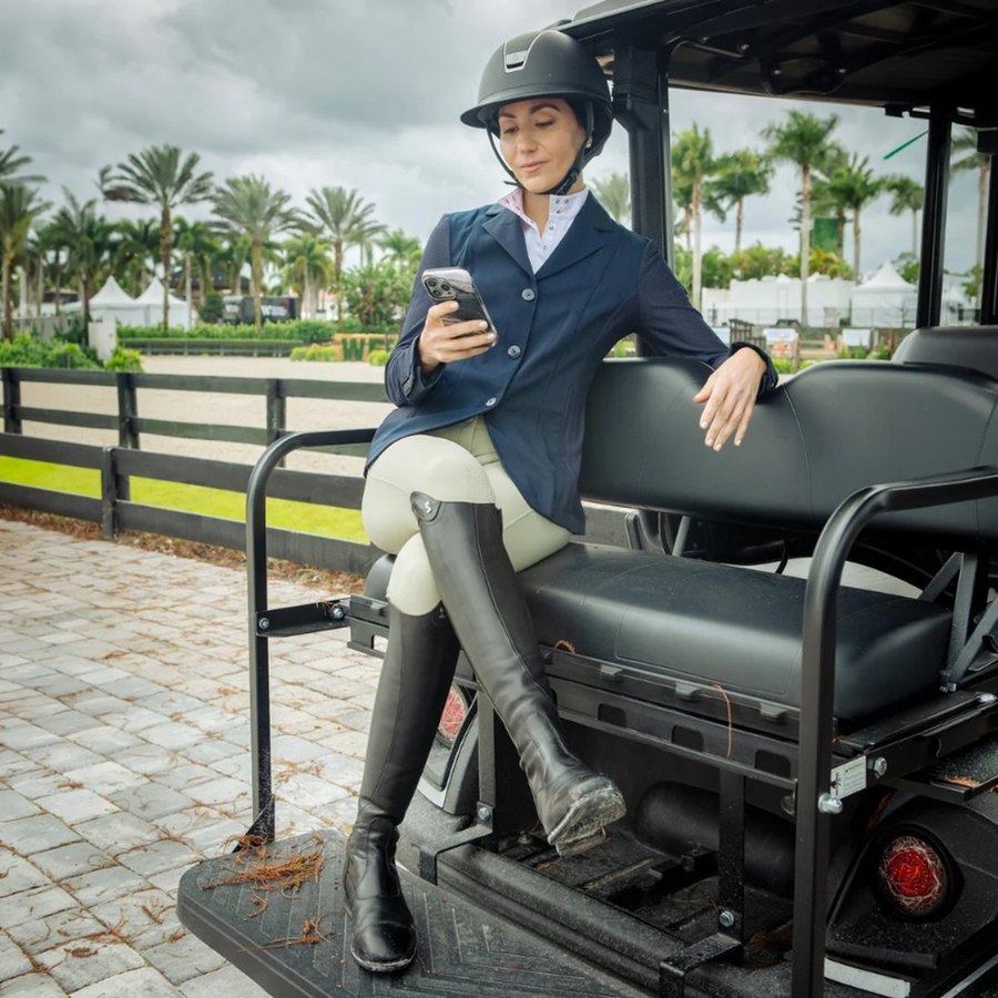 A rider in a helmet, show coat and tall boots sitting on a cart at a horse show