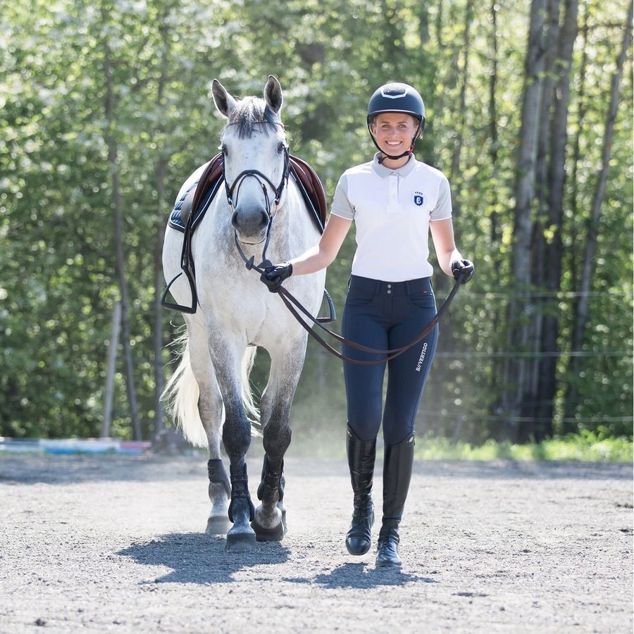 A rider in a white polo, navy breeches and tall boots leading her grey horse across the arena