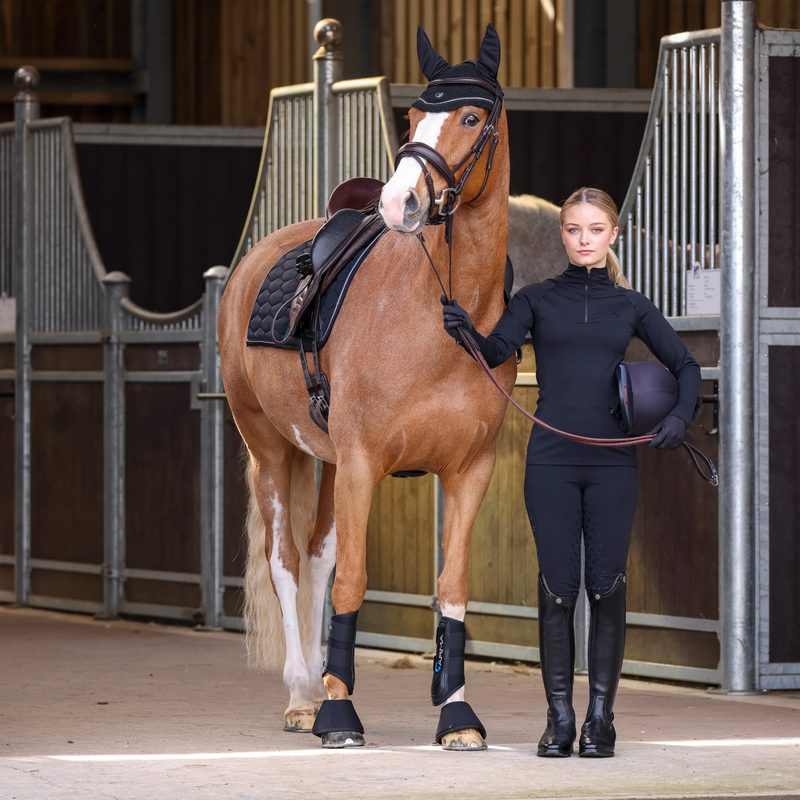 A rider in navy Shires Aubrion riding tights and tall boots standing with her chestnut horse in a barn aisle