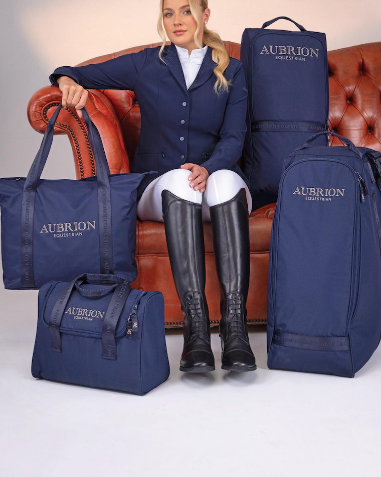 A rider in a navy show coat on a red sofa with navy Shires Aubrion Equestrian luggage around her