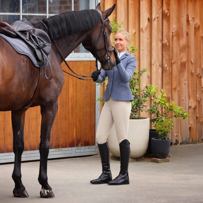 A rider in a blue Shires Aubrion show jacket, tan breeches and tall boots beside her bay horse