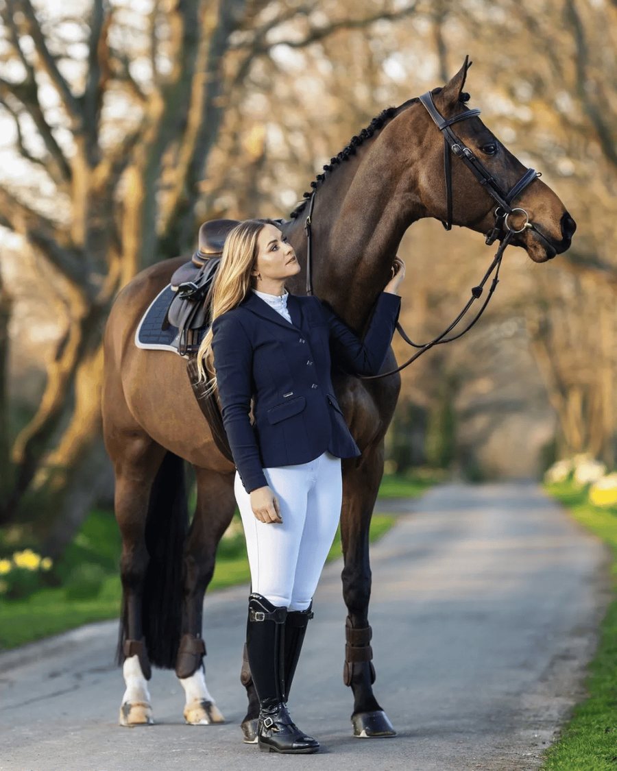 A rider in a navy show coat, white breeches and tall boots with her bay horse