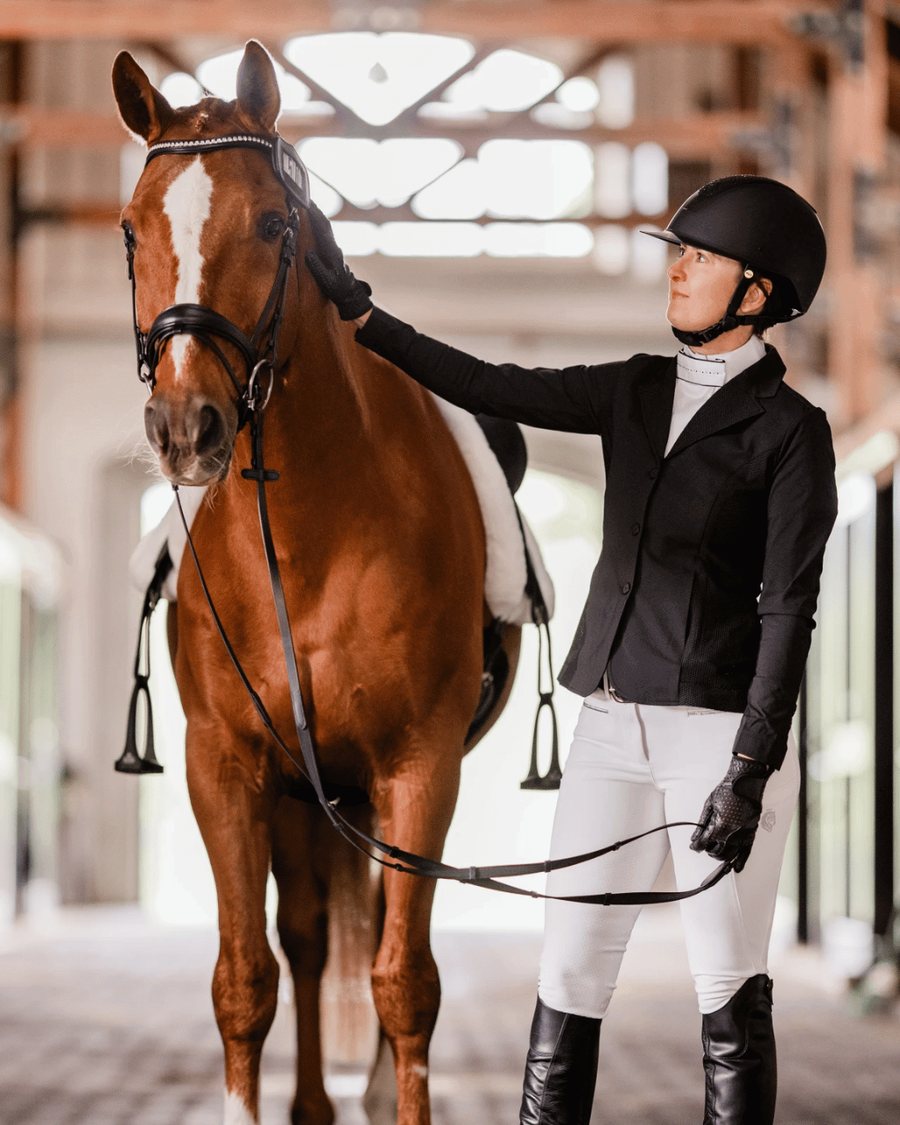 A rider in a black show coat and white breeches standing with her horse in the barn aisle