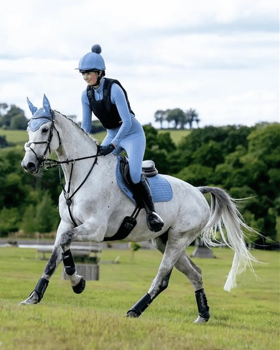 An event rider in a body protector and helmet cover cantering a gray horse across a cross-country field
